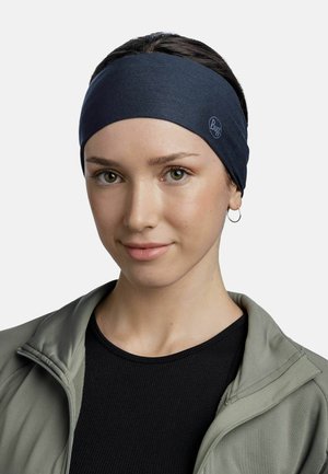 Young woman wearing a dark blue Buff headband, small hoop earring, black top, and light gray zip-up jacket, looking at the camera with a slight smile.