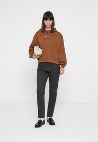 Brun cropped hettegenser med snor, paret med mørkegrå slim jeans. Modellen holder en kremfarget veske, iført svarte loafers og hvite sokker.
