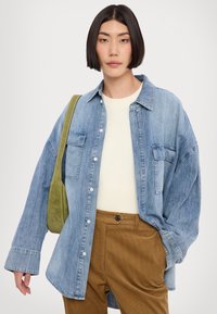 Cămașă din denim bleu deschis cu închidere cu nasturi, două buzunare pe piept și o croială lejeră. Purtați-o peste un pulover crem cu striații și pantaloni din catifea raiată maro.