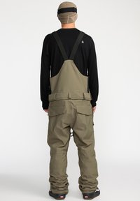 Groene geïsoleerde overalls hebben verstelbare banden, meerdere zakken en een duurzame textuur, gecombineerd met een zwart long-sleeve shirt en een beige beanie.