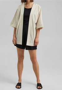 Kevyt, kerma kimonopaita pystysuorilla raidoilla; rento istuvuus, leveät hihat, avoin etuosasuunnittelu. Yhdistetty mustaan toppiin ja shortseihin.