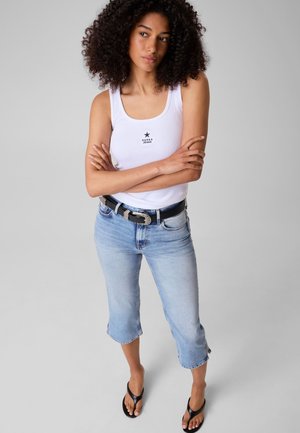 Femme aux cheveux bouclés portant un débardeur blanc, un jean bleu clair raccourci avec une ceinture noire et des tongs noires, debout, les bras croisés.