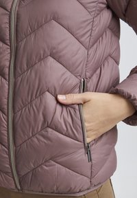 Veste matelassée à motif en chevrons dans un doux mauve. Présente une fermeture éclair centrale et des poches latérales, avec une texture lisse et une coupe ajustée.