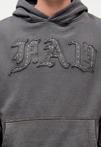 Γκρι φούτερ με κουκούλα, με τα γράμματα «MAD» σε γοτθικό στυλ, φθαρμένα, διακοσμημένα με μικρές ασημένιες τρουκς στο στήθος πάνω από την μπροστινή τσέπη.