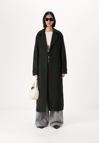 Μακρύ, σκούρο πράσινο παλτό από μαλλί, διπλό συστροφή, με μεγάλο γιακά και τσέπες. Συνδυάζεται με γκρί παντελόνι σε φαρδιά γραμμή και κρεμ τσάντα.