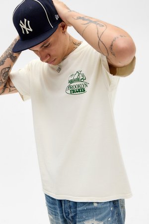 T-shirt creme com um design gráfico verde apresentando um camião e o texto "BROOKLYN FRUITS." Usado com um boné bordado em azul marinho e jeans desbotados.