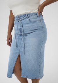 Falda de mezclilla azul claro con abertura frontal, cinturón ajustable con lazo, diseño clásico de cinco bolsillos y silueta ajustada.