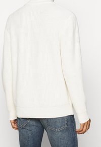 Pull blanc en maille, texture côtelée, manches longues et col montant. Assorti à un jean bleu, illustrant une tenue décontractée et confortable.