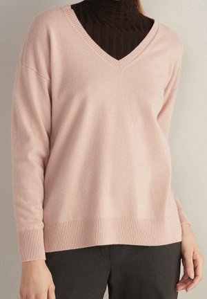 Personne portant un pull à col en V rose clair sur un col roulé côtelé foncé et un pantalon noir, debout les mains détendues le long du corps.