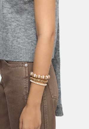 Ręka nosząca trzy bransoletki z koralików w białych i złotych tonach, zestawiona z brązowymi spodniami i szarym bezrękawnikiem z dzianiny.