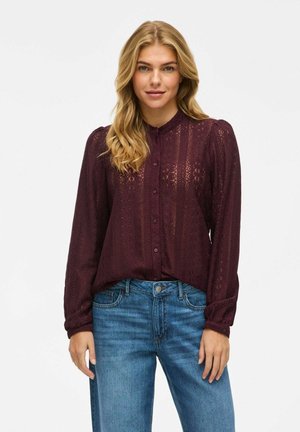 Blouse bordeaux à col montant, manches longues et texture à motifs. Associée à un jean en denim bleu clair. Boutons sur le devant.
