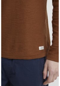 Brown knit long-sleeve sweater with a tag at the hem, featuring a fitted design and ribbed cuffs; paired with navy blue pants.