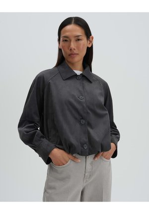 Vrouw met stijl donker haar draagt een donkergrijze jas met knopen en lichtgrijze broek, staand met handen in de zakken tegen een effen achtergrond.