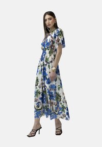 Vestido maxi floral con base blanca, que presenta patrones en azul y verde. Cintura elástica y mangas cortas; combinado con sandalias de tacón negras.