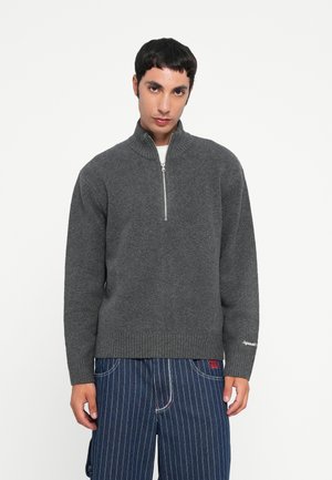 Gray knit sweater with a high collar and zippered front. Features wide ribbed cuffs and hem. Worn with dark striped fabric pants.