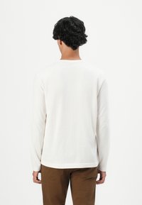 Άνδρας με σκούρα σγουρά μαλλιά, φορώντας ένα απλό λευκό πουκάμισο με μακριά μανίκια και καφέ παντελόνι, απεικονιζόμενος από πίσω σε λευκό φόντο.