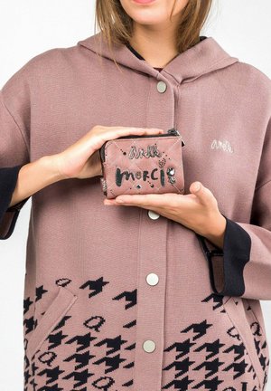 Portafoglio rosa trapuntato con accenti neri, che presenta la parola "merci" e borchie decorative; tenuto da una persona in un lungo cappotto rosa chiaro con cappuccio.