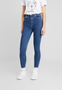 Džínové kalhoty s vysokým pasem v modré barvě, úzkého střihu, s pěti kapsami a poutky na pásek, spárované s bílým tričkem s potiskem a bílými teniskami.