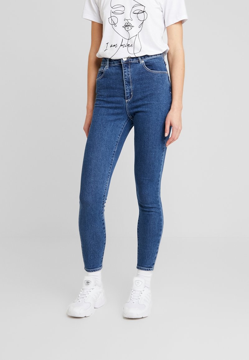 Džínové kalhoty s vysokým pasem v modré barvě, úzkého střihu, s pěti kapsami a poutky na pásek, spárované s bílým tričkem s potiskem a bílými teniskami.