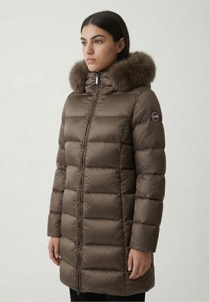 Frau trägt einen langen braunen Steppmantel mit fellbesetzter Kapuze und steht mit dem Gesicht nach links vor einem schlichten Hintergrund.