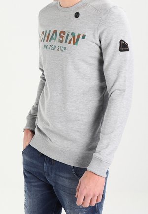 Sweat-shirt gris clair avec le texte "CHASIN' NEVER STOP" en imprimé camouflage, porté avec un jean bleu déchiré, mains dans les poches.