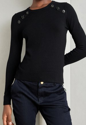 Personne portant un pull noir ajusté à manches longues avec des boutons décoratifs noirs et argentés sur les épaules et un pantalon bleu foncé.