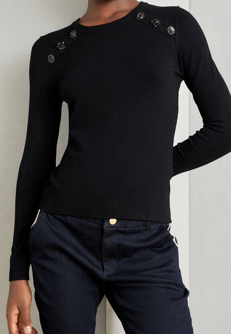 Personne portant un pull noir ajusté à manches longues avec des boutons décoratifs noirs et argentés sur les épaules et un pantalon bleu foncé.