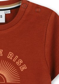 T-shirt en coton couleur rouille avec texture côtelée, présentant un col rond, deux boutons-pression et un graphique de soleil avec le texte "RISE".
