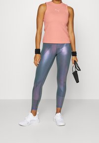 Jasnoróżowy top bez rękawów; dopasowany fason. Błyszczące turkusowe legginsy z geometrycznym wzorem. Białe trampki. Czarne opaski na nadgarstki. Skakanka w ręku.