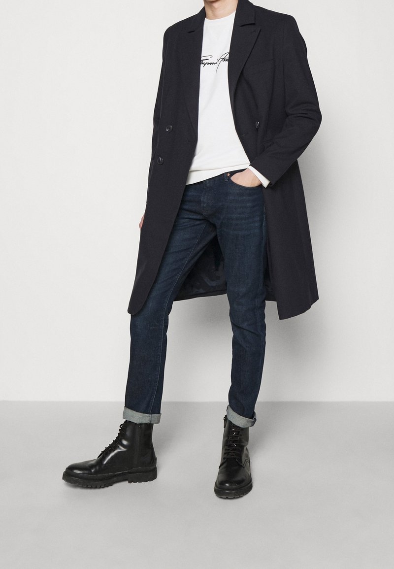 Svart dubbelknäppt kappa, vit sweatshirtd med svart text, matchat med mörka jeans uppvikta vid bensluten, och svarta snörkängor med kraftiga sulor.