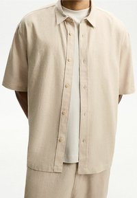 Beżowa koszula z krótkim rękawem, z zapięciem na guziki i klasycznym kołnierzykiem. Wykonana z teksturowanej tkaniny, z delikatnymi przeszyciami.