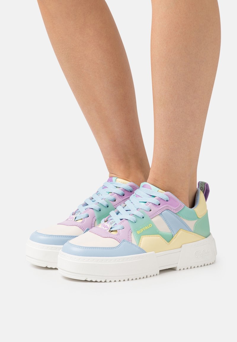 Personne portant des baskets compensées aux couleurs pastel avec des panneaux bleus, violets, verts et jaunes et des semelles épaisses blanches sur un fond uni.