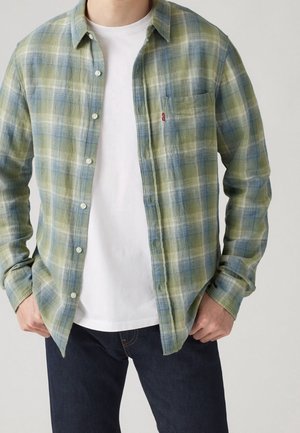 Homme portant une chemise à carreaux verte et bleue avec boutons sur un t-shirt blanc, associée à un jean bleu foncé, les mains reposant près des poches.