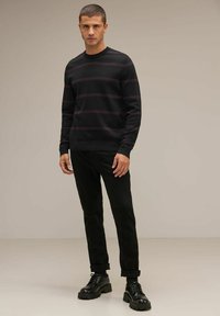 Svart stickad tröja med vinröda horisontella ränder, rund halsringning, matchad med svarta byxor och kraftiga svarta skor, mot en neutral bakgrund.