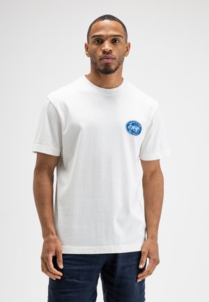 Man staand, draagt een wit T-shirt met een klein blauw rond logo op de linkerborst en donkere broek tegen een effen witte achtergrond.