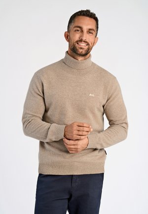 Man glimlachend, draagt een beige coltrui en donkere broek, staand met gevouwen handen tegen een effen lichte achtergrond.