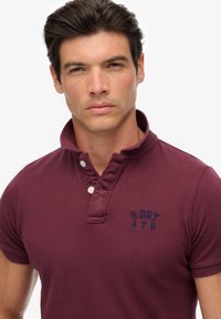 Bordeaux polohemd van een katoenmix met een zachte kraag, twee witte knopen en een marineblauw geborduurd logo op de borst. Korte mouwen.