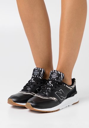 Beine mit schwarzen New Balance 997H Sneakern, die weiße Zebramuster auf Zunge und Kragen, weiße Zwischensohlen und gummi-farbene Außensohlen haben.