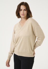 Pulover bej cu decolteu în V dintr-un material moale, tricotat, cu cusături pe umeri căzute și manșete cu pliuri, asortat cu blugi negri.