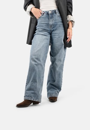 Une personne portant un jean bleu taille haute, un haut blanc et un blazer noir, se tenant avec une main dans sa poche et des bottes marron.