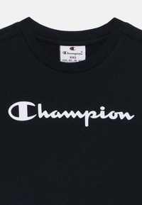 Juodas medvilnės marškinėlis su baltu "Champion" logotipu kursyvu priekyje. Apvalus kaklas ir etiketė matoma viduje.