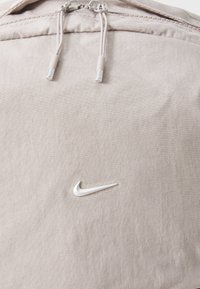 Nike-laukku vaaleassa beigen kankaassa, jossa on sileä pinta, hopeiset yksityiskohdat ja iso valkoinen Swoosh-logo edessä.