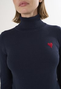 Maglione a collo alto blu navy realizzato in materiale morbido, con un piccolo cuore rosso ricamato sulla parte sinistra del petto.