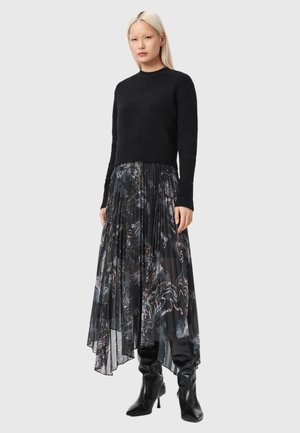 Frau mit langen blonden Haaren, die einen schwarzen Pullover, einen plissierten gemusterten Midirock und schwarze Stiefel mit hohen Absätzen trägt, steht vor einem grauen Hintergrund.