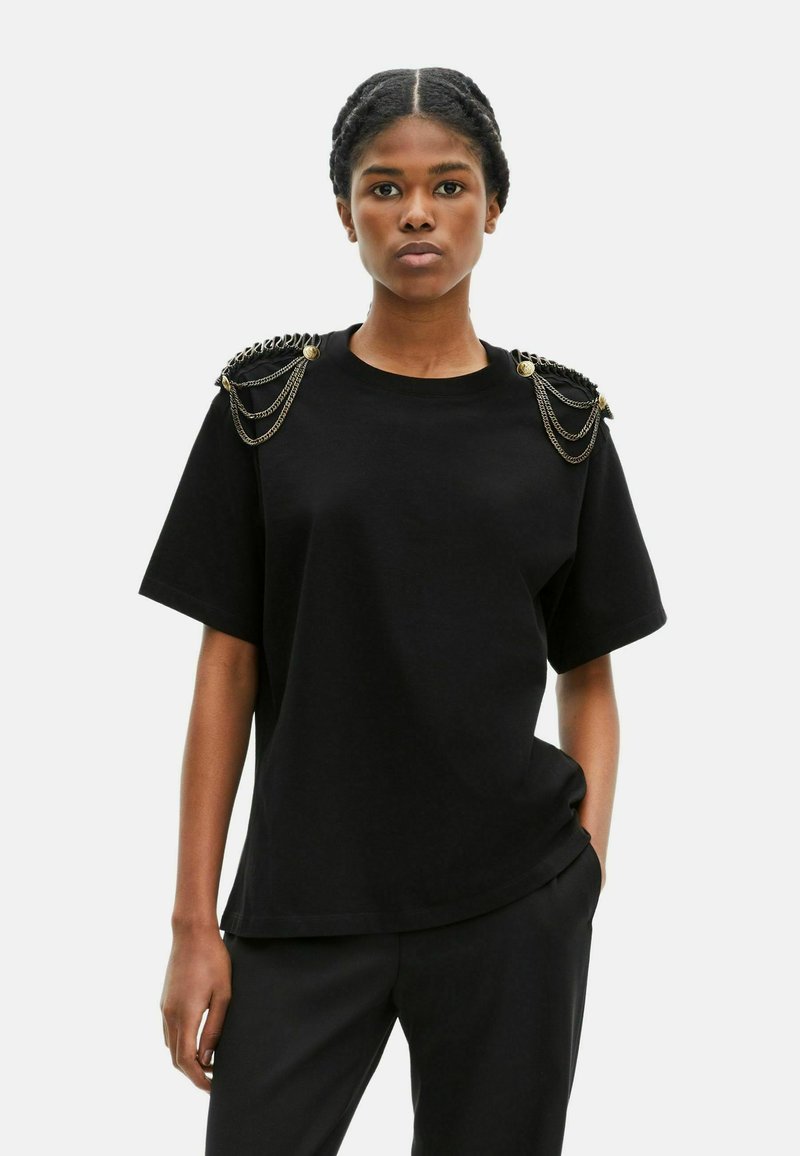Camiseta de algodón negra de manga corta, con acentos de cadena metálica en los hombros y un corte relajado. Textura suave, diseño minimalista.
