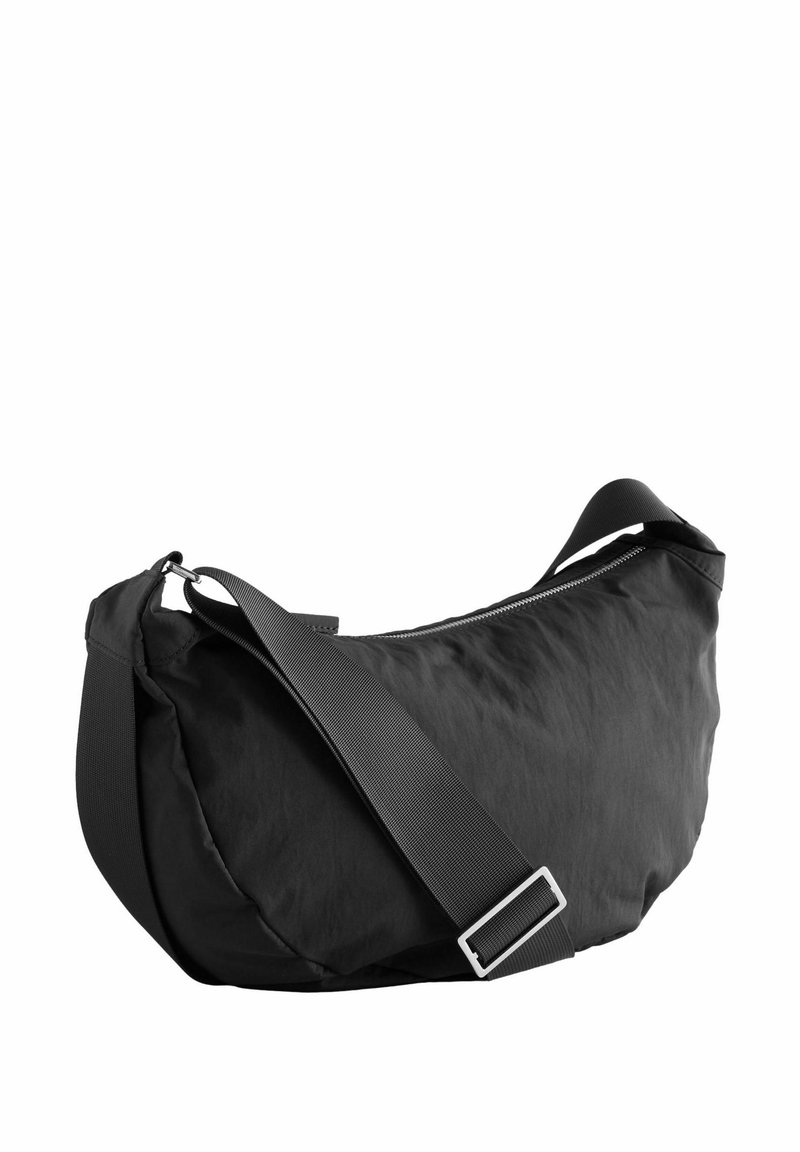Bolso de hombro de tela negra con forma curva, correa de nylon ajustable y cierre superior con cremallera. Textura suave con acentos de hardware mínimos.