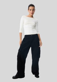 Weiße Langarmbluse kombiniert mit schwarzen weit geschnittenen Cargo-Hosen, die über Seitentaschen und einen Knopfverschluss verfügen; glatte Textur.