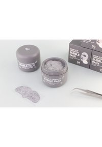 Un pot gris étiqueté "Pack de Bulles Carbonatées en Argile Colorée" avec un couvercle texturé, rempli d'un produit duveteux gris. Un masque gris et des pinces se trouvent à proximité.