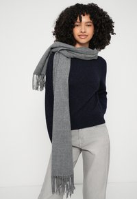 Siv šal s resicami, položen čez temno modro pulover. Mehka tekstura, pravokotna oblika in lahka zasnova. Priložnostna kombinacija z hlačami.