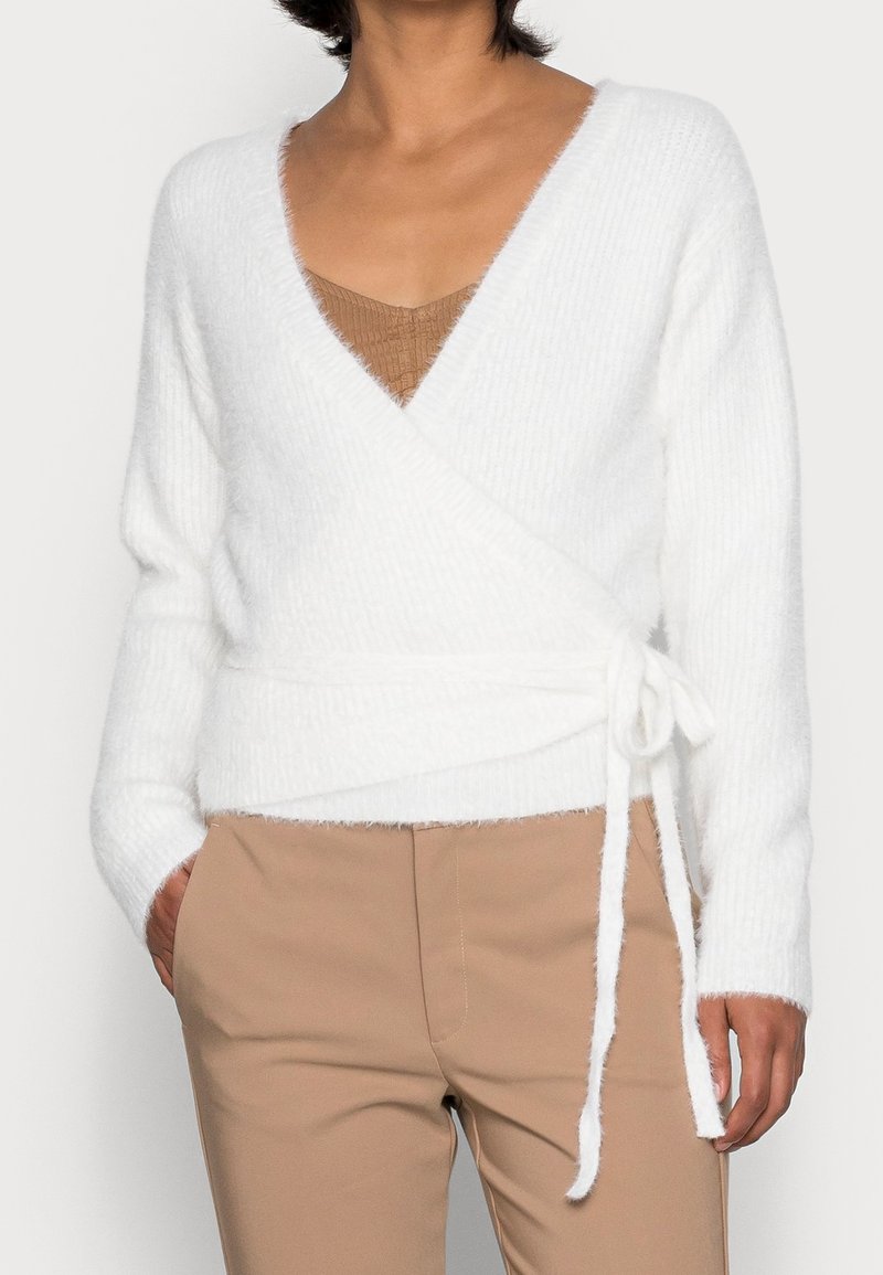 Witte, fluffy wikkeltrui met een diepe V-hals en strik in de taille; heeft lange mouwen en een geribde textuur, gelaagd over een tan top.